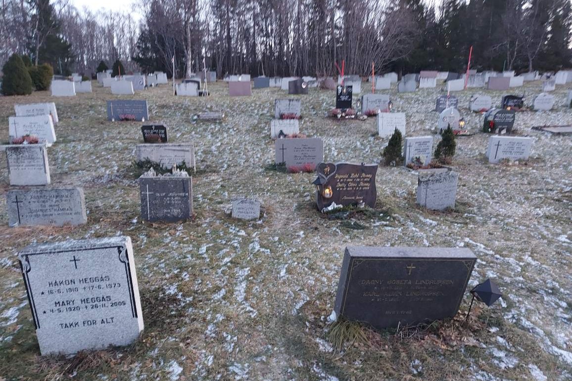Snødekt gravplass med flere gravminner, noen av dem stående litt skjevt på gressbakken.
